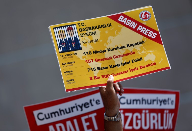 A raised hand holds a large ID card showing journalists in jail in place of a photo.